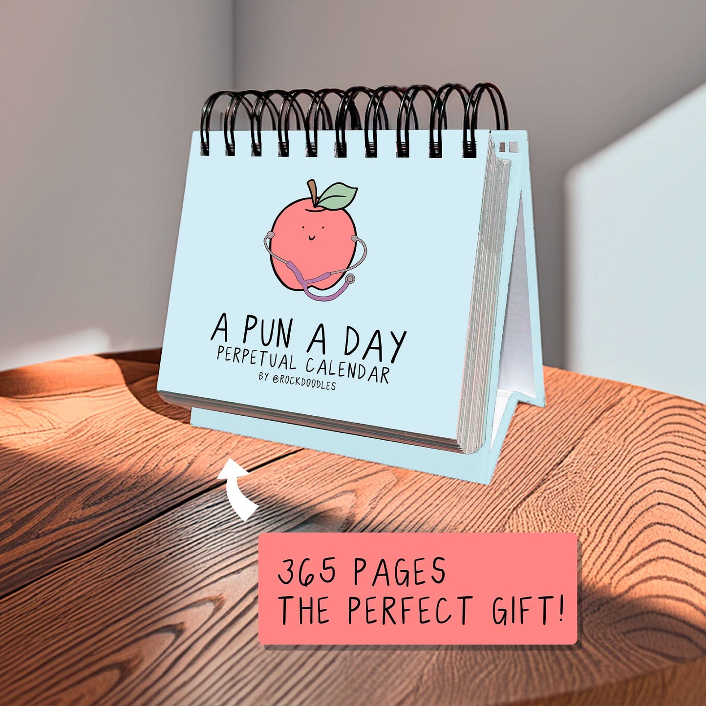 The "A Pun A Day Perpetual Calendar" by rockdoodles, featuring a cartoon peach on its spiral-bound cover, sits on a sunlit wooden table. Note: “365 pages of daily puns—the ideal wordplay gift!”.