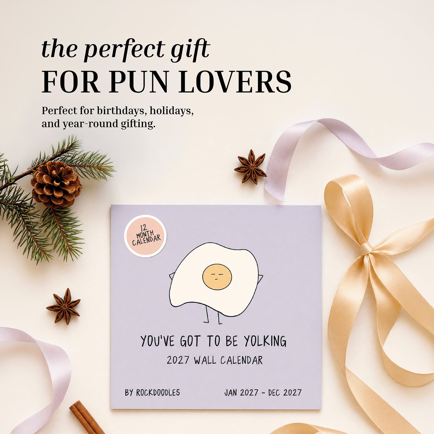 The rockdoodles 2027 Wall Calendar, "You've Got To Be Yolking," showcases a playful egg cartoon and witty pun, set among pinecones, ribbon, star anise, and a gold gift bow on a light surface. This funny wall calendar makes an ideal gift for any pun enthusiast.