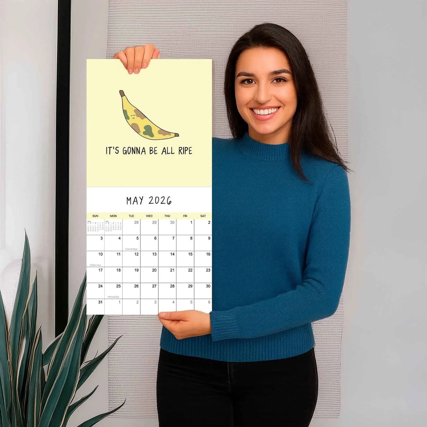 A woman in a blue sweater holds up the 2026 wall calendar "You've Got To Be Yolking" by rockdoodles, showing May's page with a cartoon banana and the text "It's gonna be all ripe.