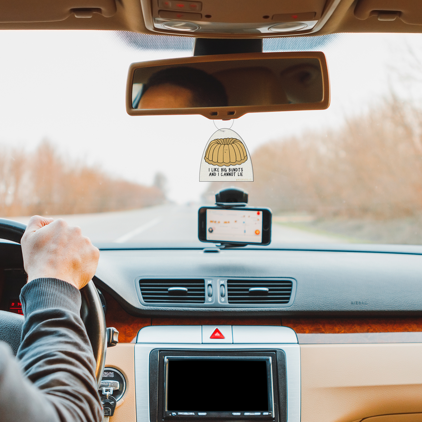 A man driving a car with two rockdoodles Big Bundt (2 Pack) Punny Air Fresheners - Vanilla Scent on the dashboard.