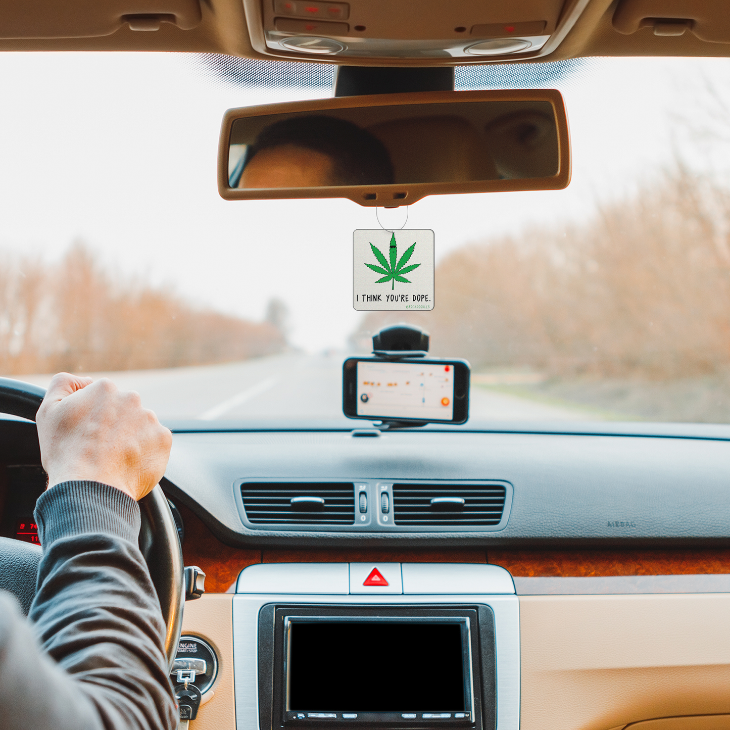 A man driving a car with a "You're Dope (2-Pack) Punny Air Freshener - Pine Forest Scent" hanging from the dashboard, made by rockdoodles.