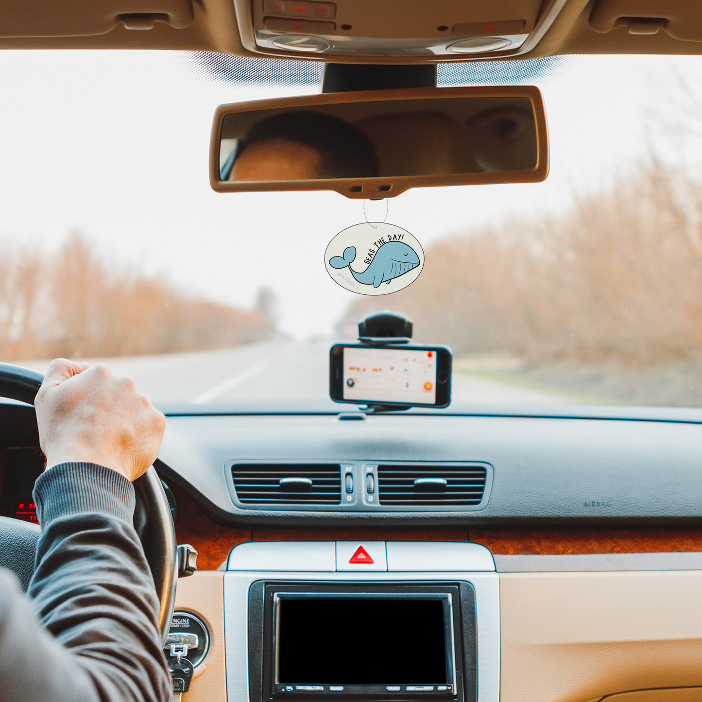 A man driving a car with a Seas The Day (2-Pack) Punny Air Freshener - Ocean Scent, by rockdoodles, on the dashboard.