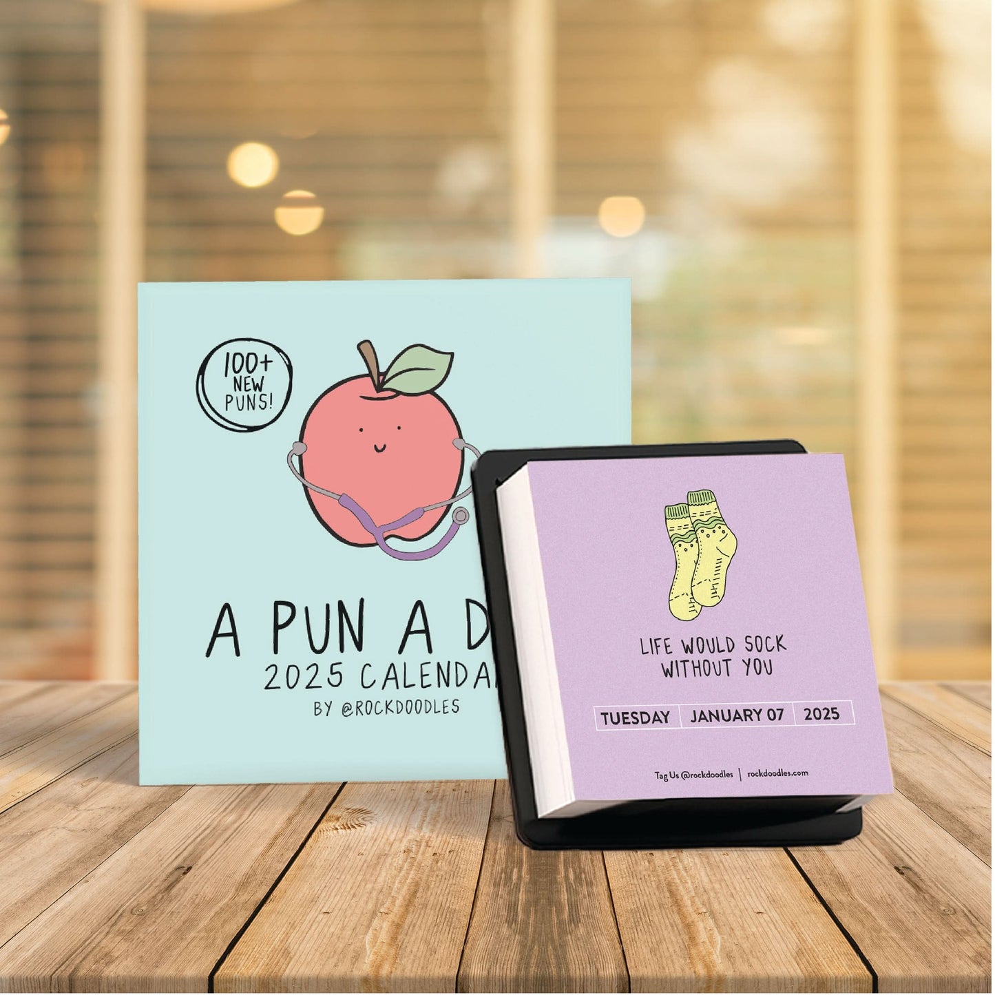 Two 2025 A Pun A Day Desktop Calendars by rockdoodles with whimsical illustrations—a peach on one, yellow socks and a January 7, 2025 page on the other—are shown on wood. Ideal as an inspirational desk calendar.