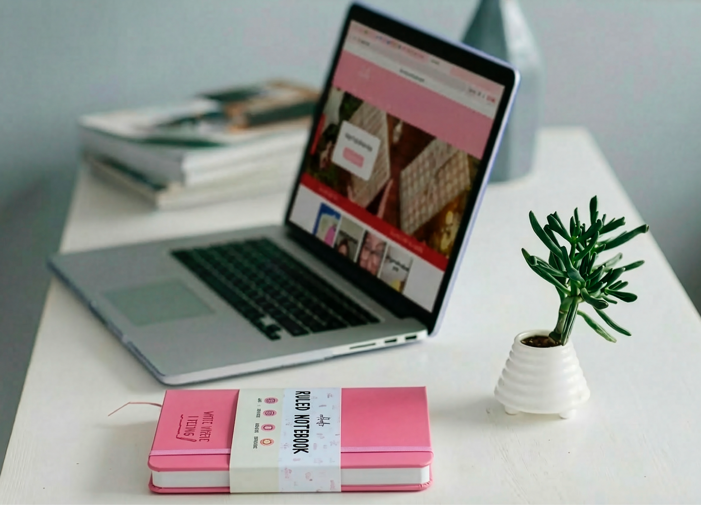 A laptop with an open website sits on a white desk next to a small plant, a creative journal, two stacked Rockdoodles Notebooks from rockdoodles—one college ruled—and some magazines.