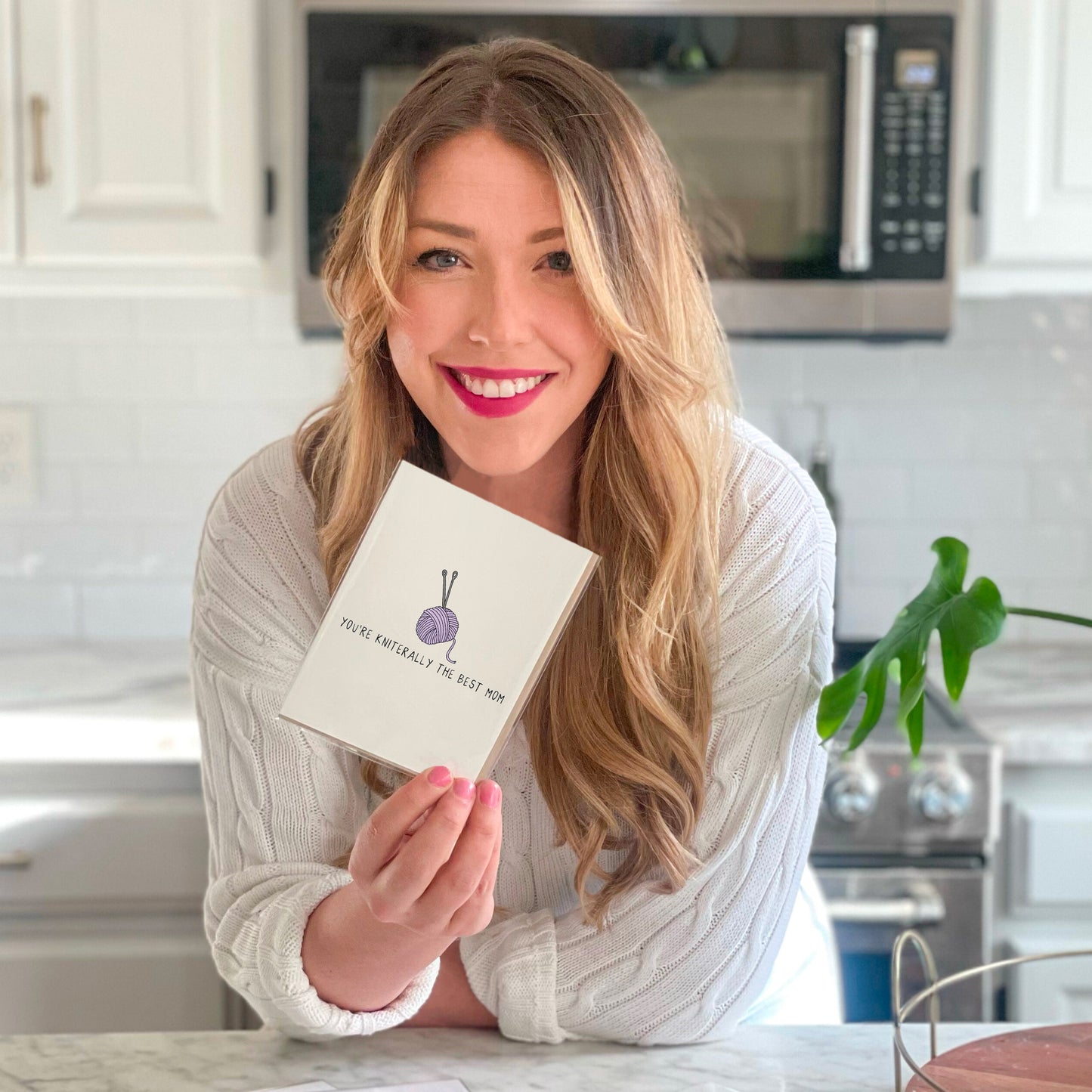 A woman in a kitchen holding up the rockdoodles You're Kniteraly The Best Mom Card.