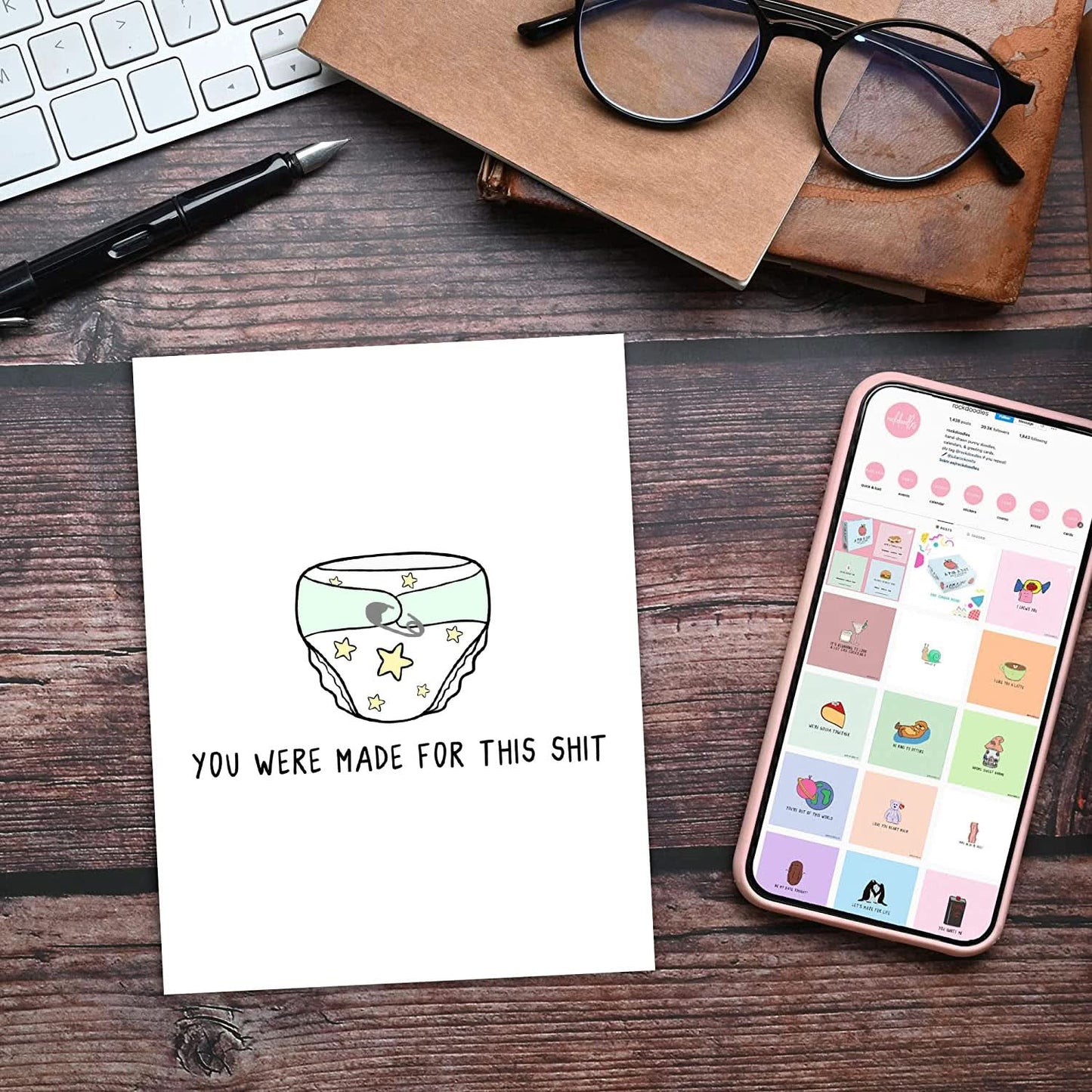 A greeting card with a diaper illustration and the text "You Were Made For This Shit" lies on a wooden desk, alongside a smartphone, a pair of glasses, a notebook, a pen, and a keyboard. The Made For This Shit Card by rockdoodles is BLANK INSIDE and comes with a natural envelope.