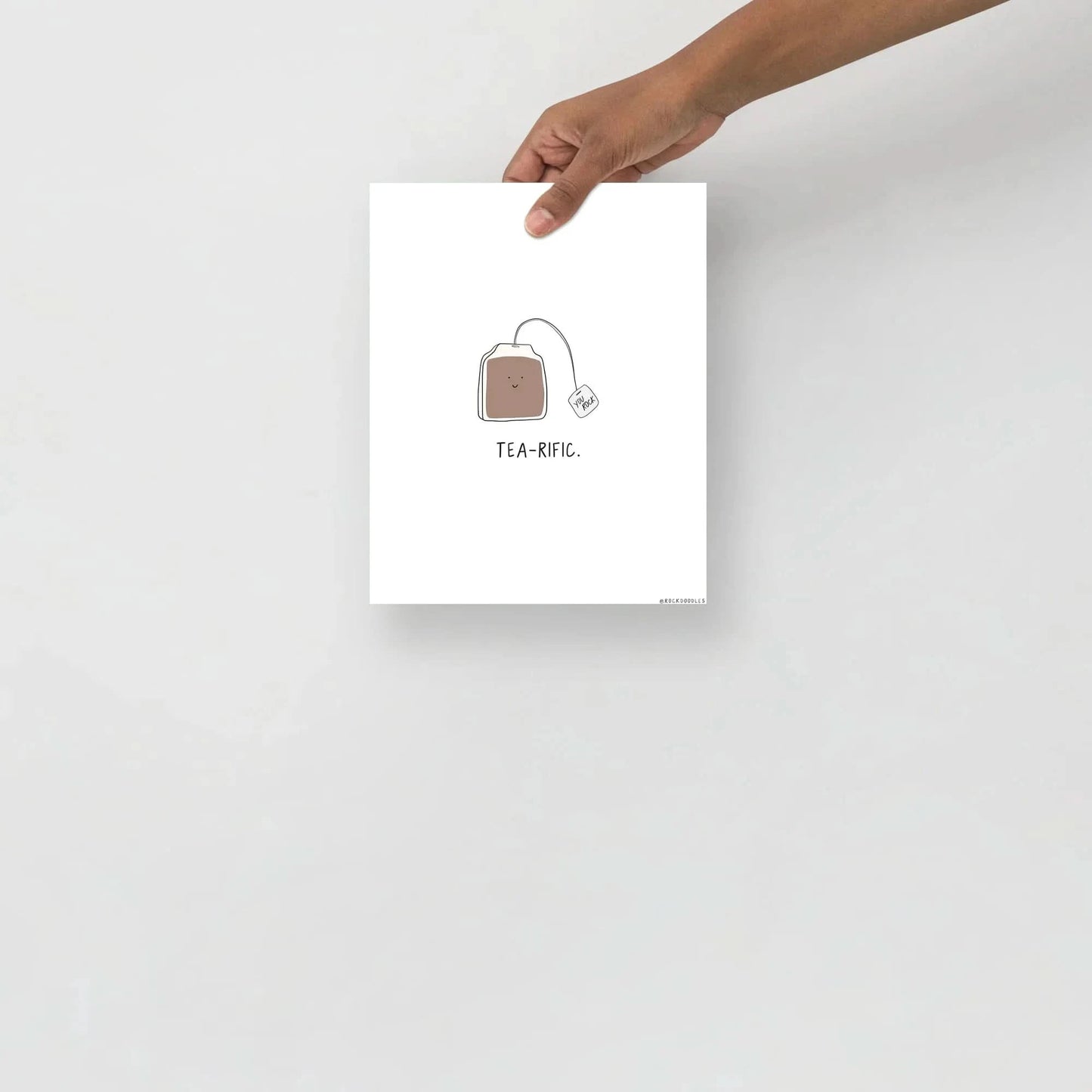 A person holding up a "Tea-rific Print" poster from the brand rockdoodles, printed on thick matte paper sourced from renewable forests.