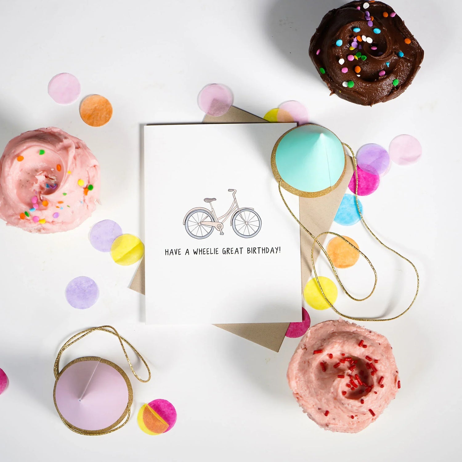 A birthday card with a bicycle illustration and the text "Have a wheelie great birthday!" is surrounded by three cupcakes, confetti, and two gift tags on a white surface.