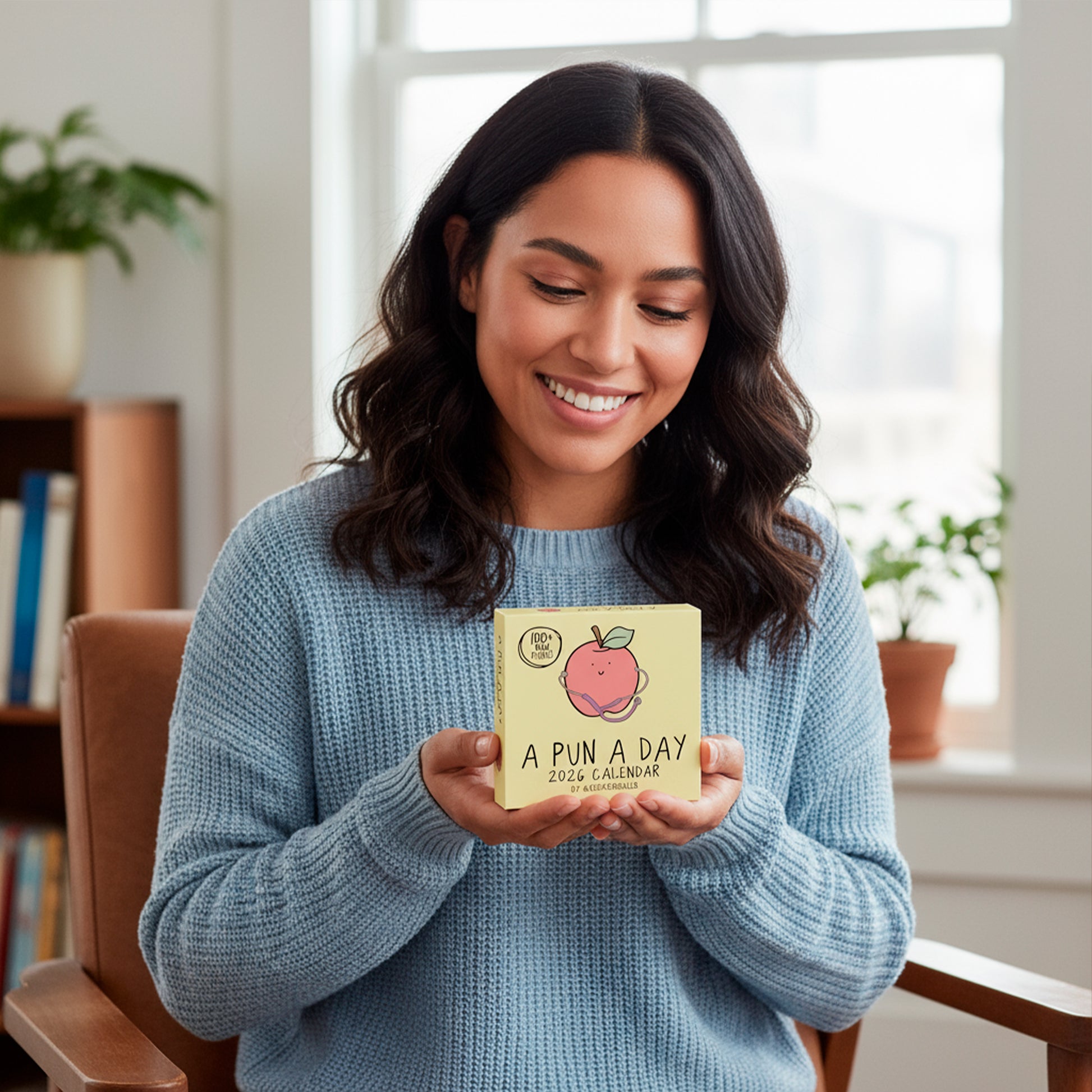 A woman in a blue sweater sits indoors, smiling and holding a small 2026 Calendar Gift Set by rockdoodles—perfect for pun lovers or anyone searching for fun, unique gifts.