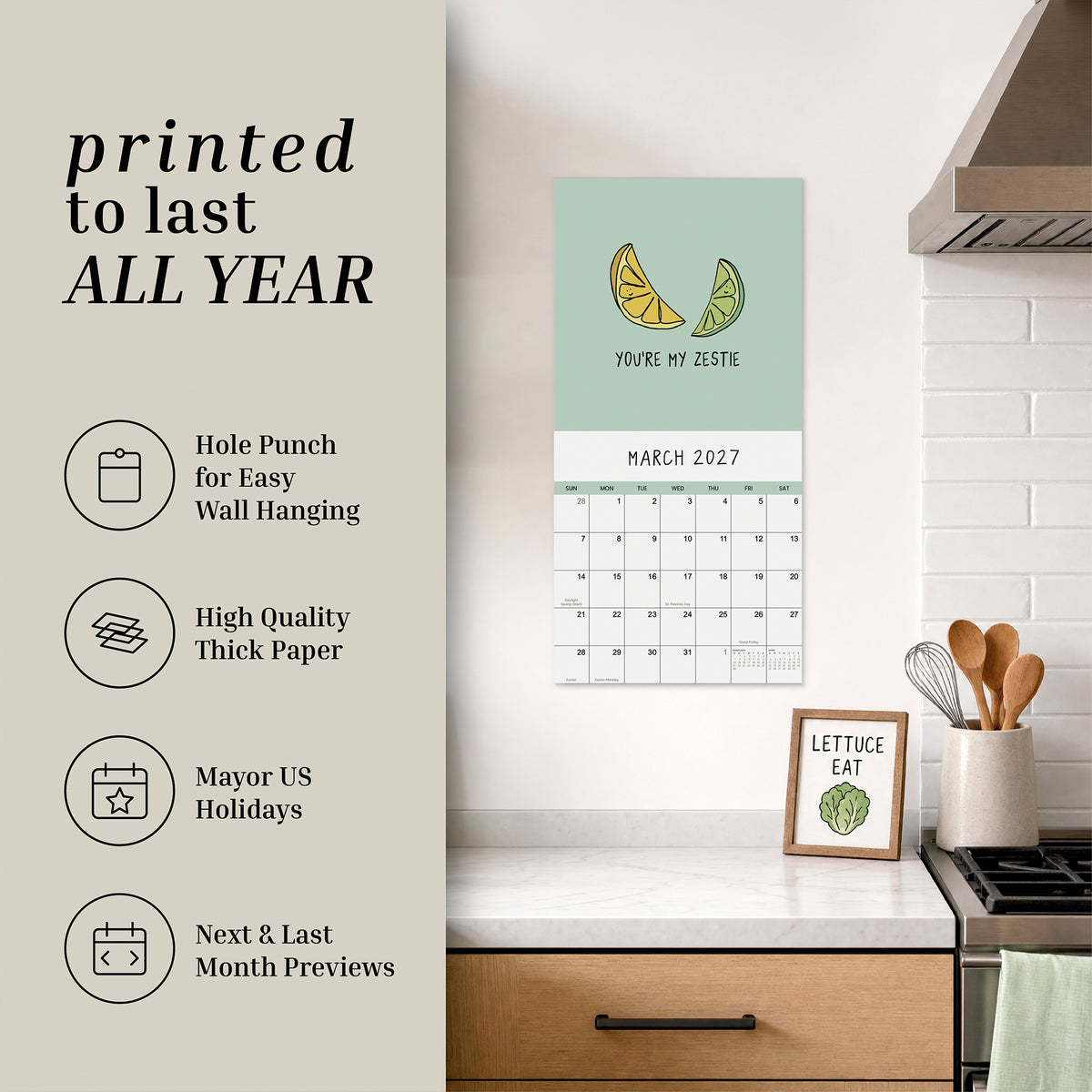 A 2027 wall calendar from rockdoodles titled "You've Got To Be Yolking" hangs above a kitchen counter adorned with decor, kitchen tools, and a framed drawing of lettuce—bringing a touch of whimsy to the space with its pun-filled calendar charm.