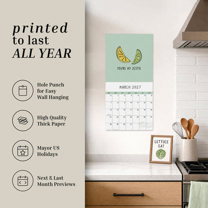 A 2027 wall calendar from rockdoodles titled "You've Got To Be Yolking" hangs above a kitchen counter adorned with decor, kitchen tools, and a framed drawing of lettuce—bringing a touch of whimsy to the space with its pun-filled calendar charm.