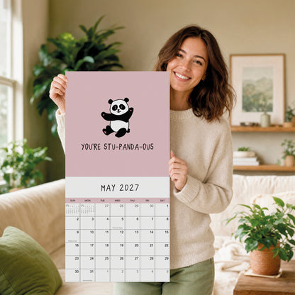 A woman smiles while holding a rockdoodles 2027 Wall Calendar—titled "You've Got To Be Yolking"—a pun calendar, in a cozy, plant-filled living room.