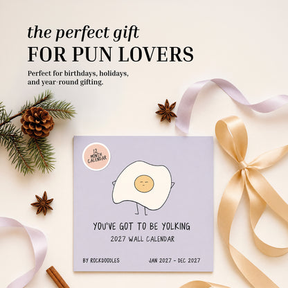 The rockdoodles 2027 Wall Calendar, "You've Got To Be Yolking," showcases a playful egg cartoon and witty pun, set among pinecones, ribbon, star anise, and a gold gift bow on a light surface. This funny wall calendar makes an ideal gift for any pun enthusiast.
