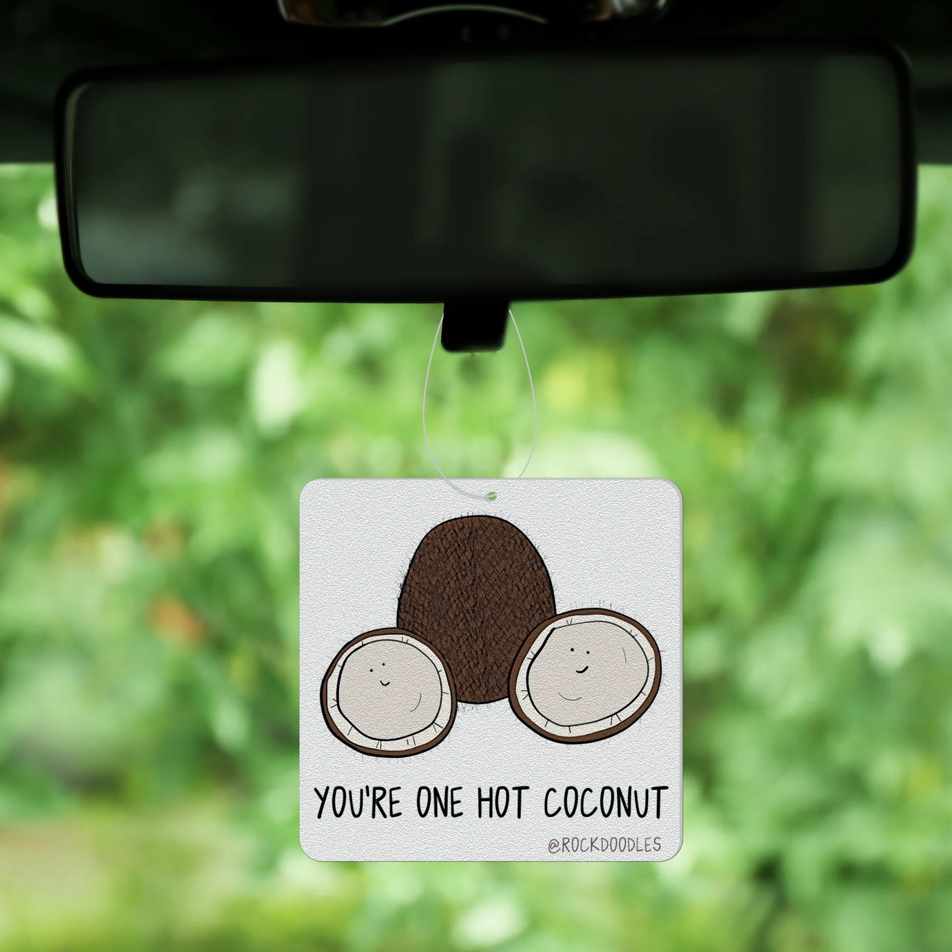 A car air freshener designed by rockdoodles, featuring two cartoon coconuts and the playful phrase "You're one hot coconut," hangs from a rearview mirror, perfect for pun enthusiasts, set against a blurred green background.