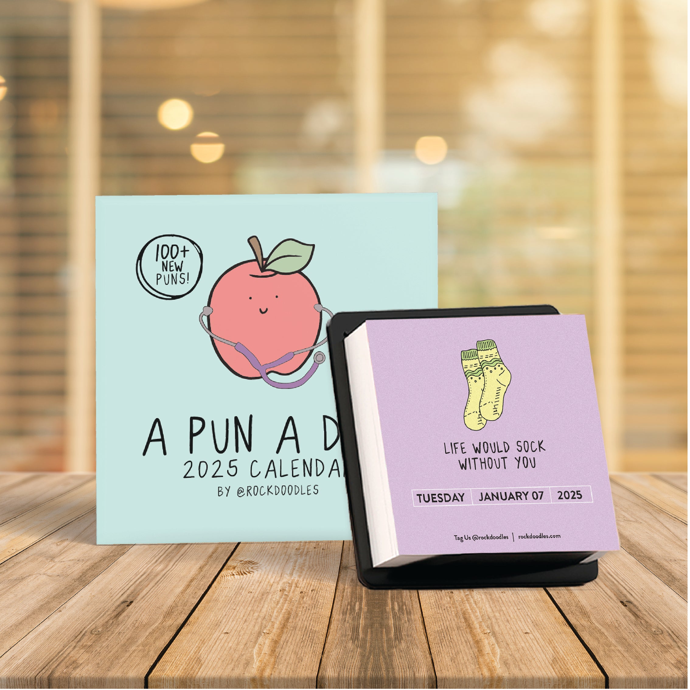 Image of two calendars on a wooden table. One features the 2025 A Pun A Day Desktop Calendar by rockdoodles, adorned with a peach pun. The other daily calendar, also from rockdoodles, showcases a sock pun, 