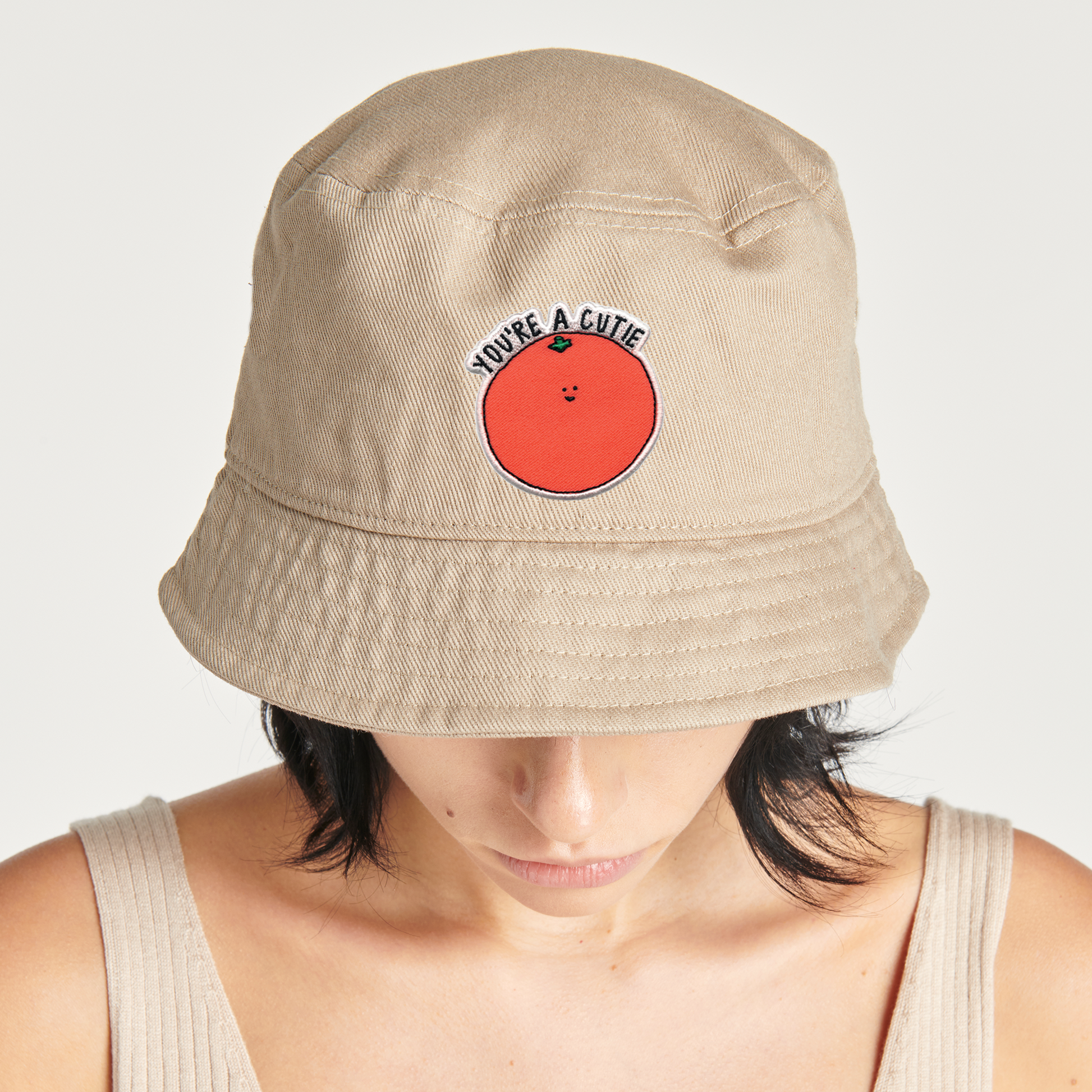 A woman wearing a tan bucket hat with a rockdoodles You're A Cutie Patch on it.