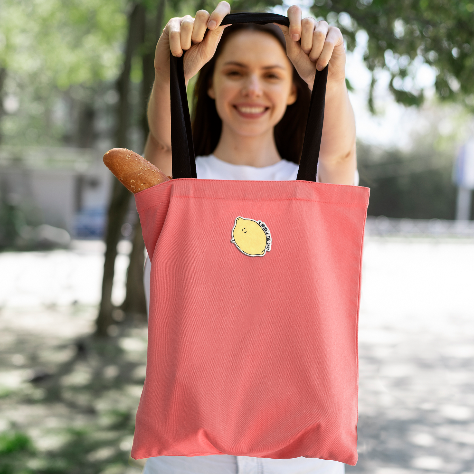 A woman holding a pink tote bag with a Squeeze The Day Patch by rockdoodles on it while following iron-on instructions using a household iron.