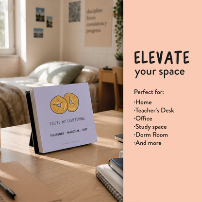The 2027 A Pun A Day Desktop Calendar by rockdoodles is featured on a wooden table in a cozy, sunlit room. Described as perfect for the home, teacher’s desk, office, study, or dorm room, this plastic-free calendar showcases a pun-filled drawing on each page—offering both eco-friendly style and delightful charm to any environment.