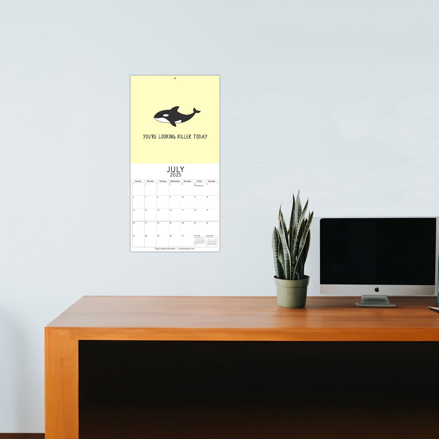 A vibrant rockdoodles calendar titled "You've Got To Be Yolking," displaying July 2025 and complete with a whale illustration and the pun-filled message "You're looking killer today," hangs above a wooden desk with a potted plant and a computer monitor.