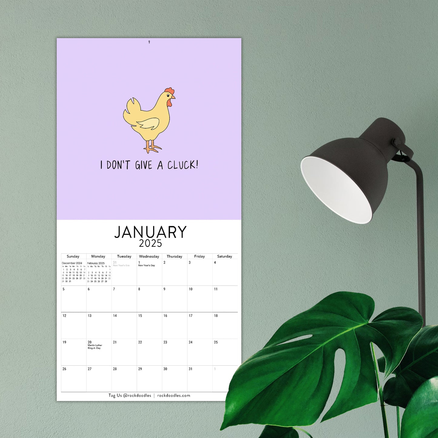 A 2025 ADULT Wall Calendar "Knotty By Nature" by rockdoodles featuring an illustration of a chicken with the cheeky humor text "I DON’T GIVE A CLUCK!" hangs on a green wall next to a black lamp and a green plant.