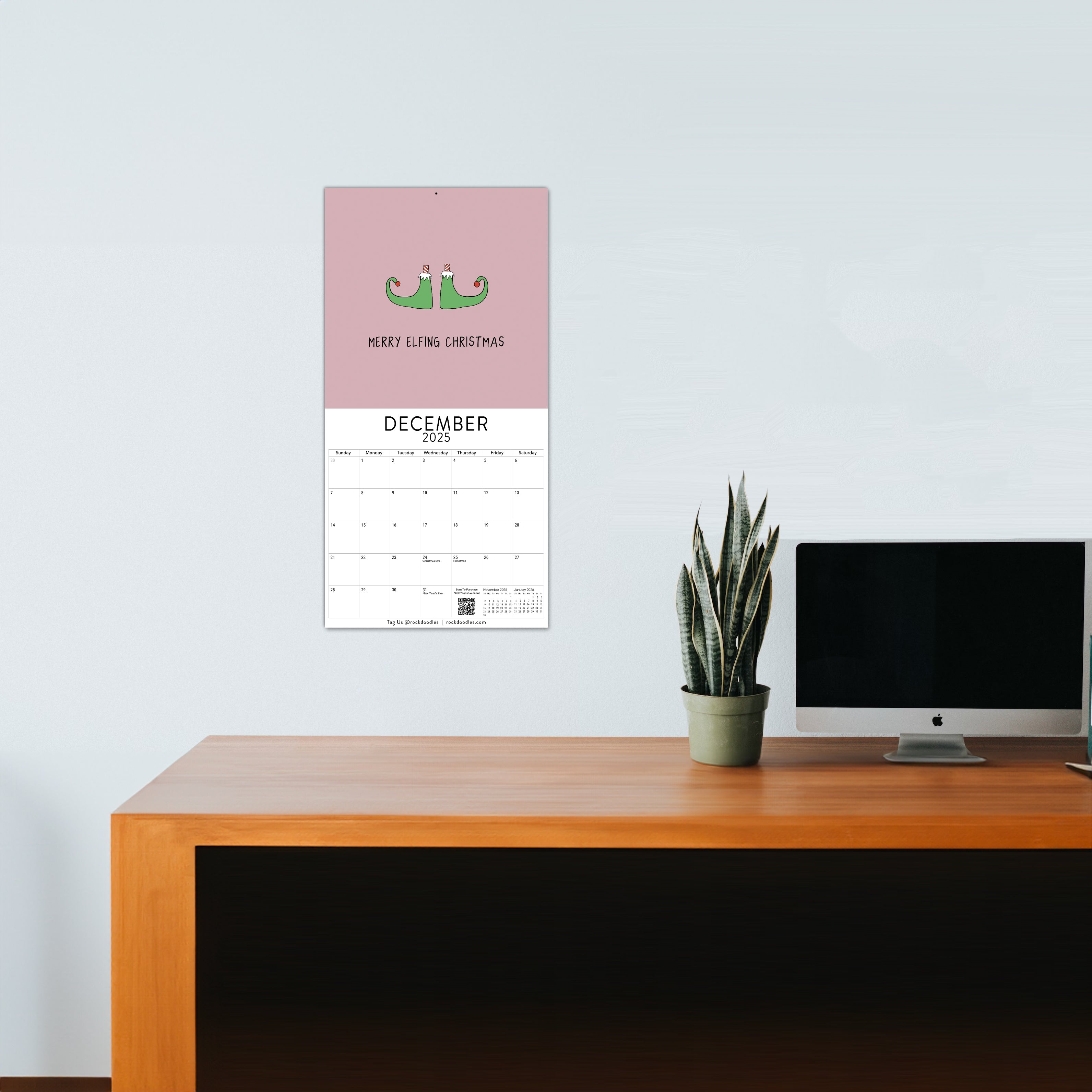 A wooden desk with a computer monitor and a potted plant; above the desk hangs the 2025 ADULT Wall Calendar 