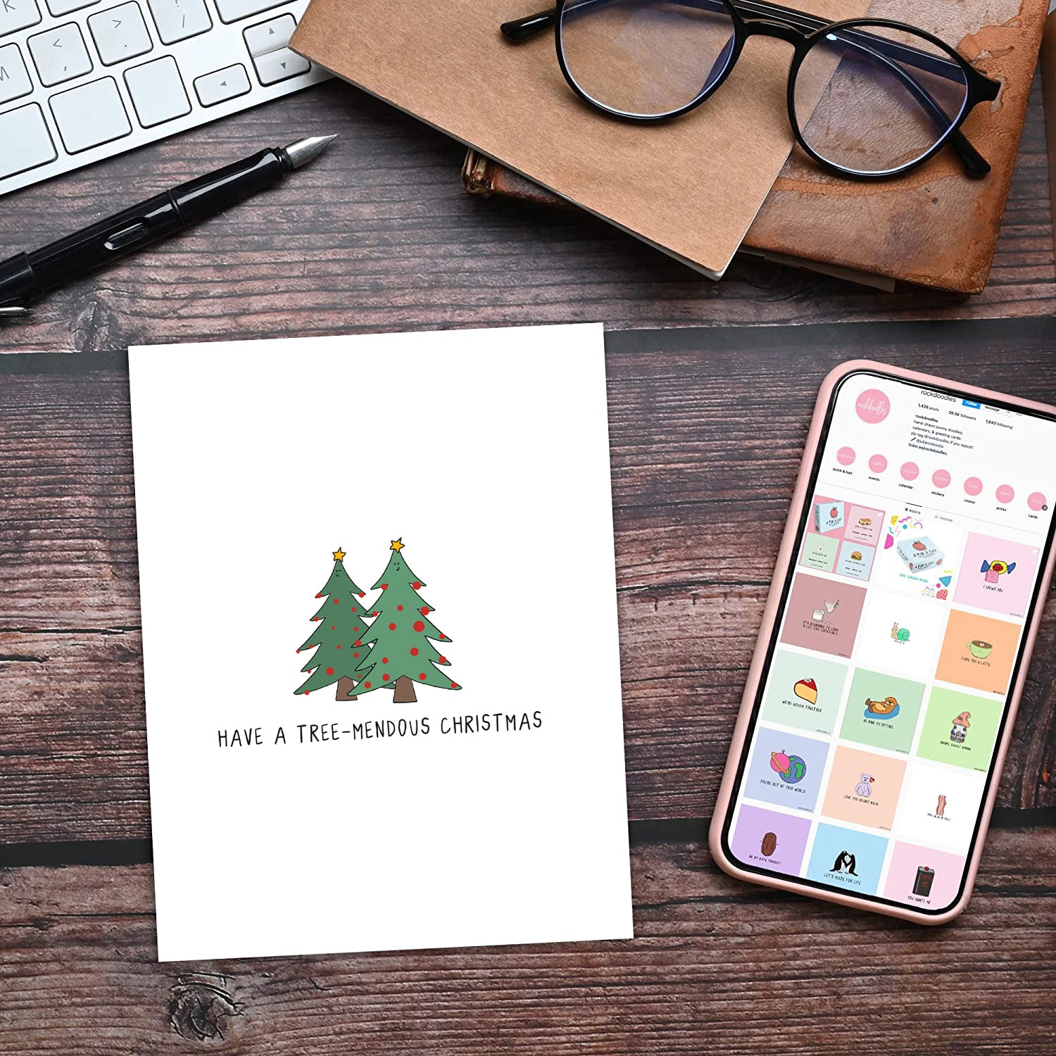 A rockdoodles Have A Tree-Mendous Christmas Card on a desk next to a phone and glasses.