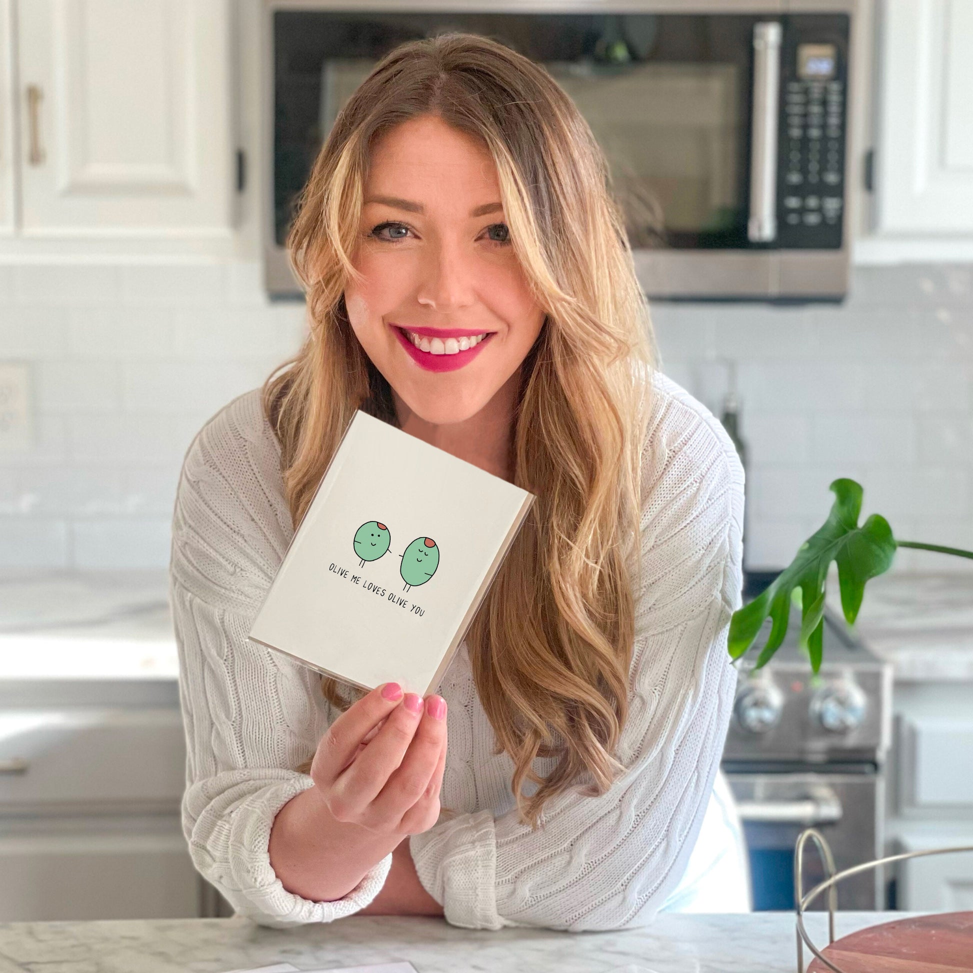 A woman is holding an Olive Me Card in front of a kitchen counter.