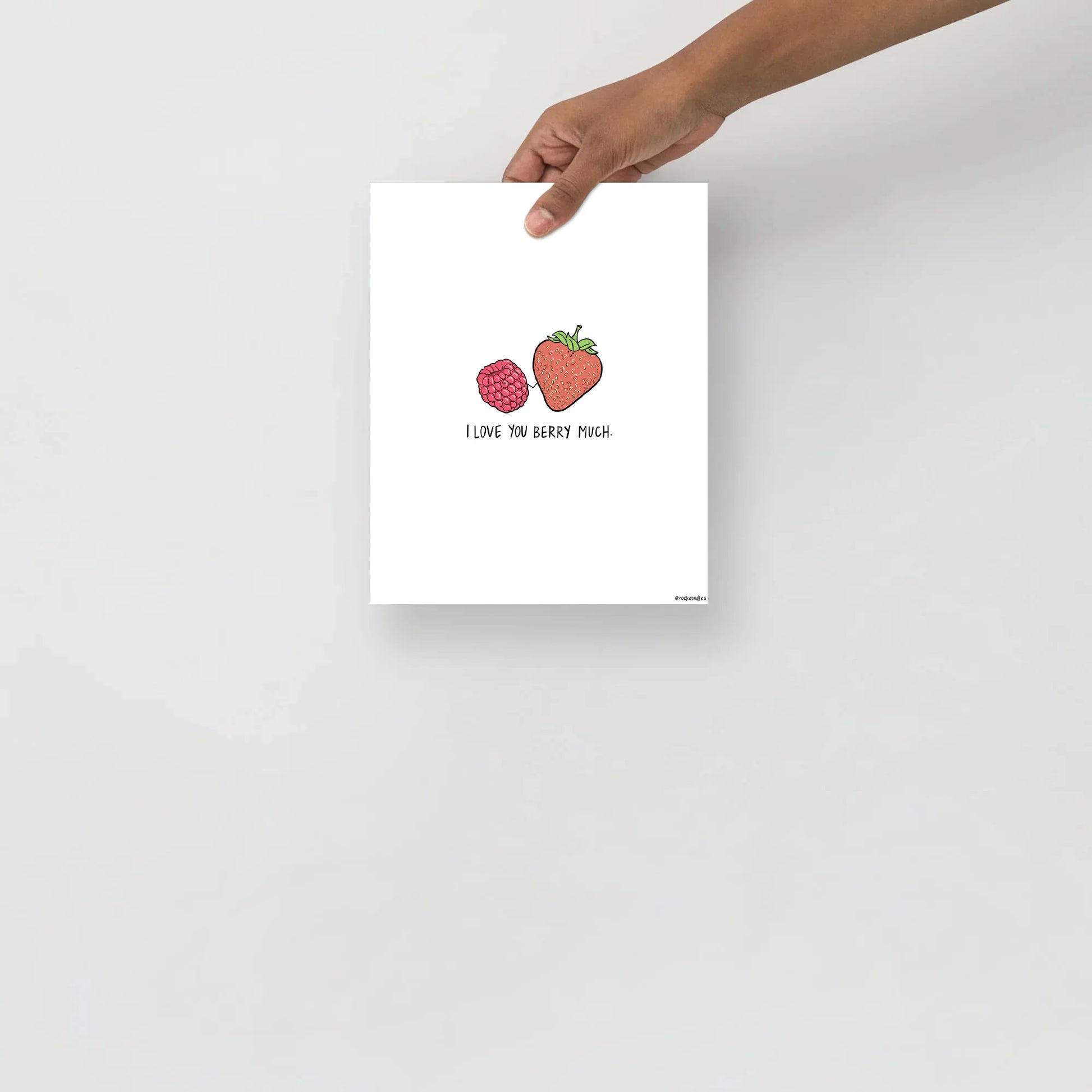 A "Berry Much Print" by rockdoodles, featuring a hand holding up a card with a strawberry on it, displayed in a wood frame.